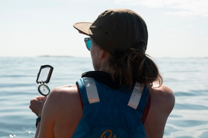 Person in a kayak holding a compass, wearing blue life vest and cap, facing open water.