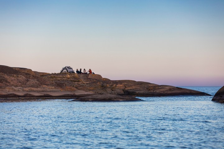 People sitting by a tent on rocky shore near calm water at sunset.