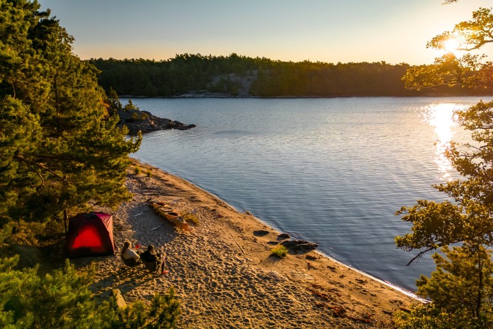 Tent and kayaks on sandy beach by forested lake at sunset.