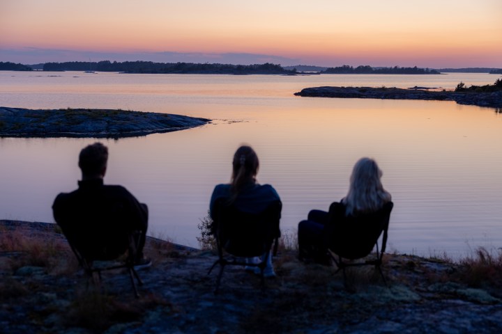 Three people seated, watching a sunset over a calm lake with distant islands.