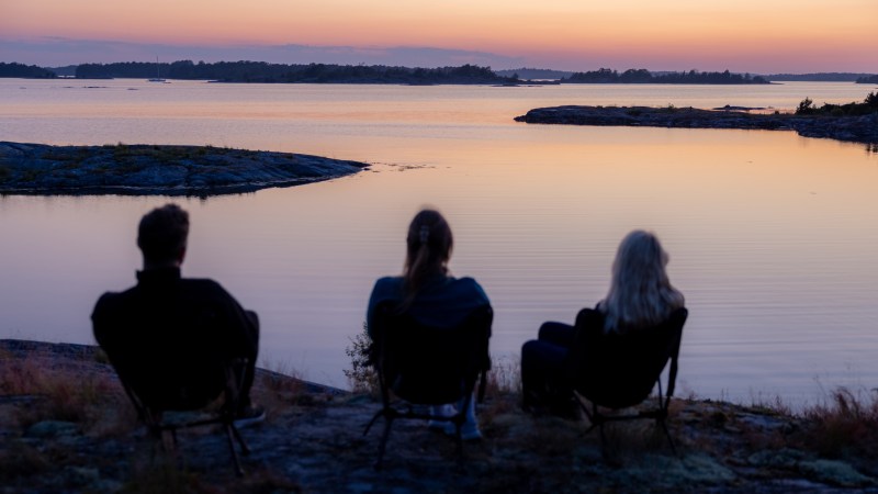 Three people seated, watching a sunset over a calm lake with distant islands.