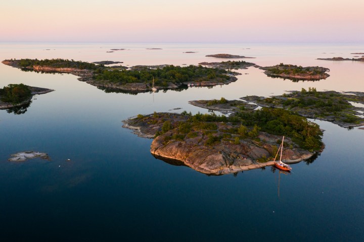 Aerial view of small islands with green trees and a sailboat on calm water under a pink sky.