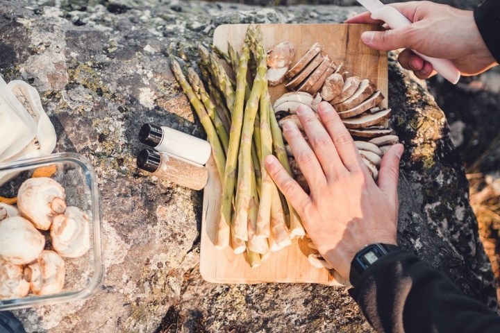 Hands slicing mushrooms and preparing asparagus on a wooden board outdoors.