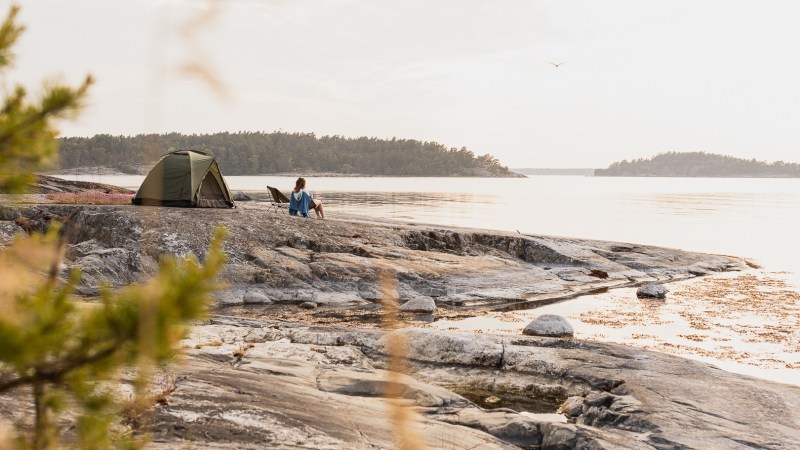 Person sitting by a tent on rocky shore, overlooking water and distant forested islands in calm weather.