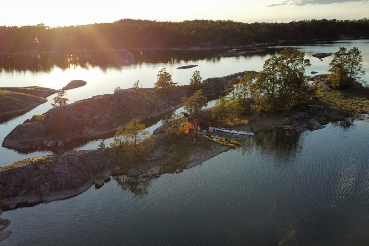 Sunset over a small island with trees, tents, and kayaks on calm water.