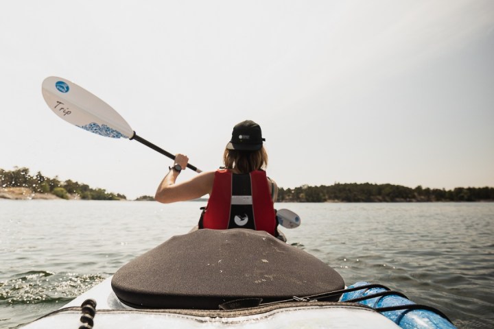Person kayaking on a calm lake, wearing a red vest and cap, with trees in the background.
