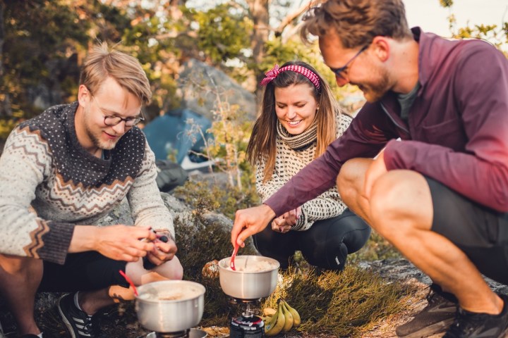 Three people cooking with camping gear outdoors, smiling and enjoying the activity under sunlight.