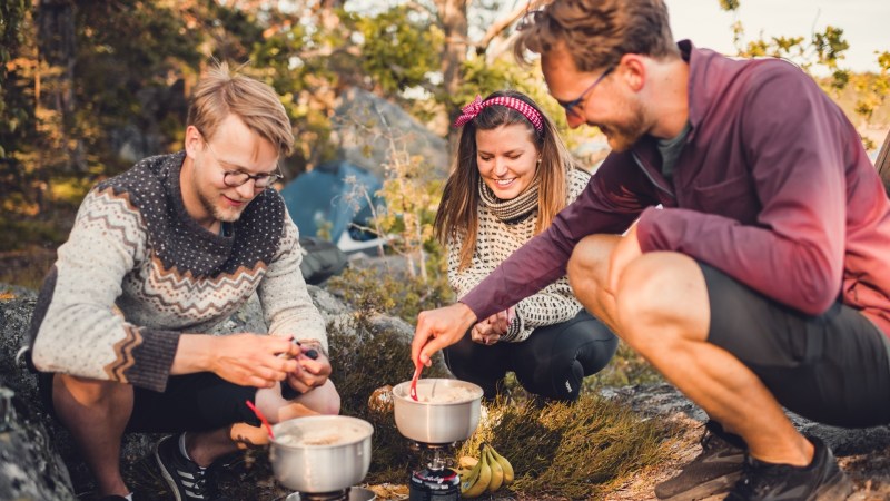 Three people cooking with camping gear outdoors, smiling and enjoying the activity under sunlight.