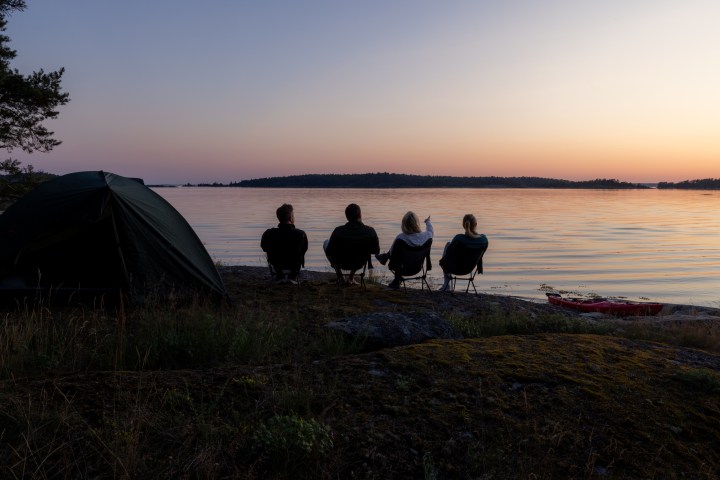 Four people seated by a tent watching a sunset over a calm lake.