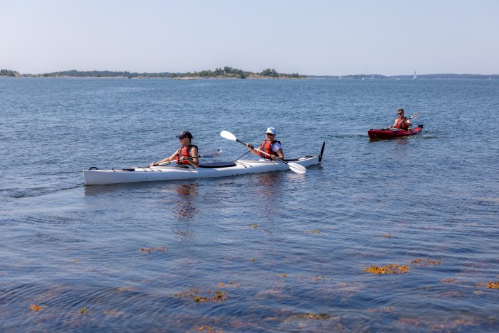 Three people kayaking on a calm sea near an island under a clear sky.