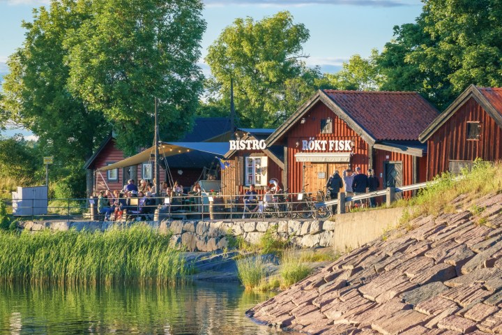 Wooden bistro by the water with people dining, surrounded by greenery and a cobblestone path.