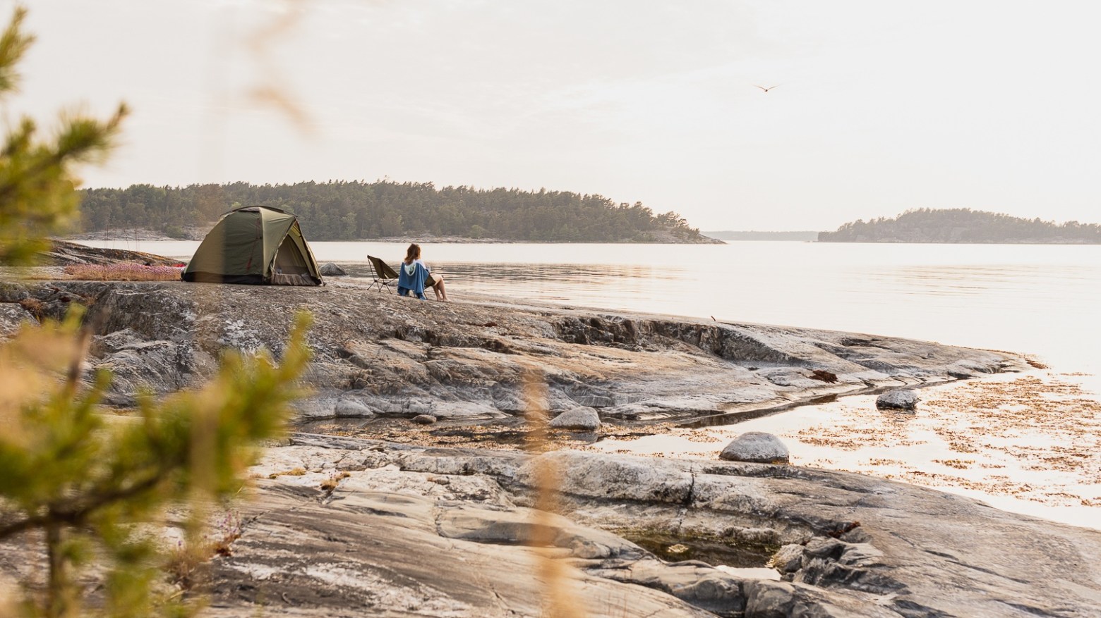 Person sitting by a tent on rocky shore overlooking calm water and distant islands.