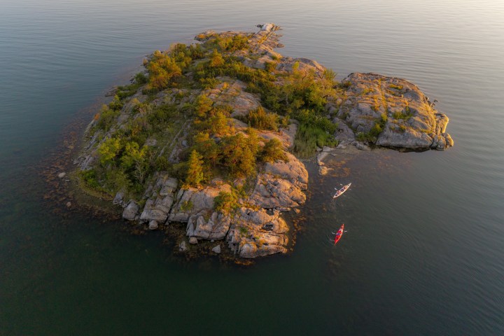 Aerial view of a rocky island with trees and two kayaks in calm water at sunset.