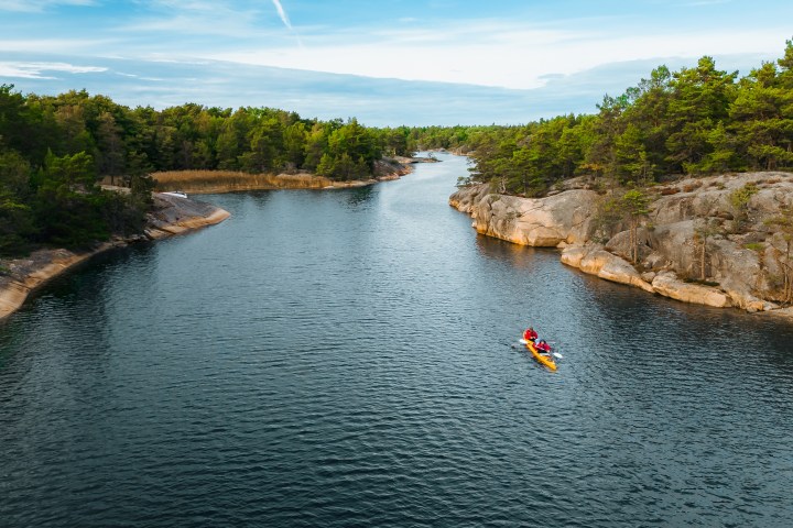 Person kayaking on a narrow waterway between rocky, forested shores under a blue sky.