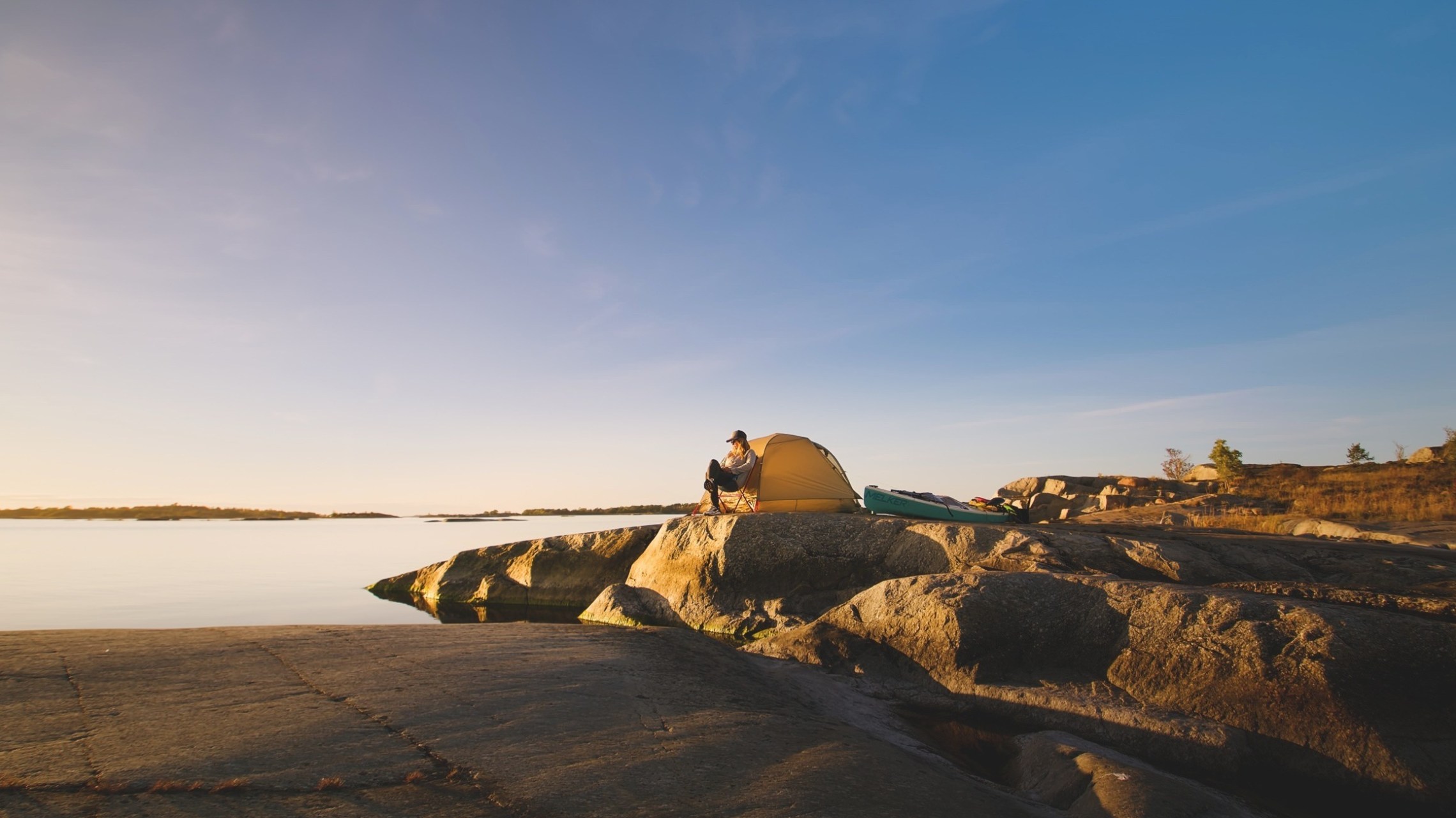 Person camping on a rocky shoreline with a yellow tent at sunset.