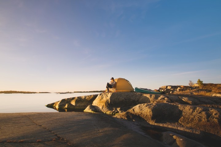 Person camping on a rocky shoreline with a yellow tent at sunset.