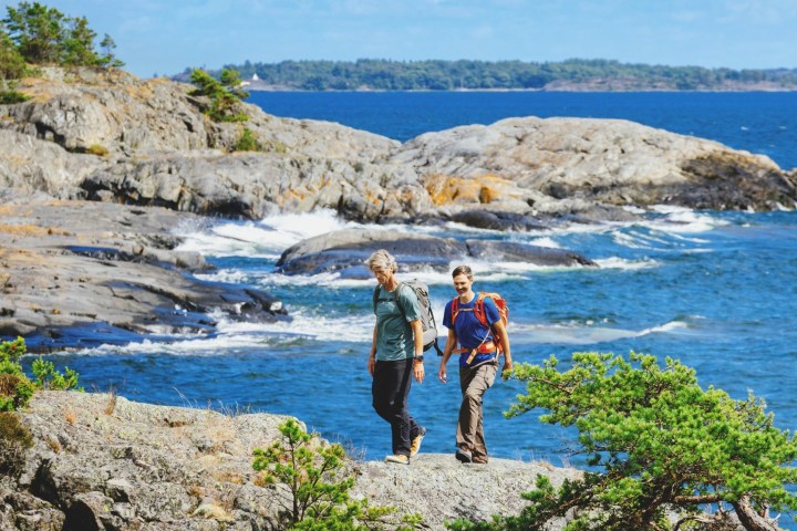 Two hikers walking on rocky coast with ocean and forested islands in background.