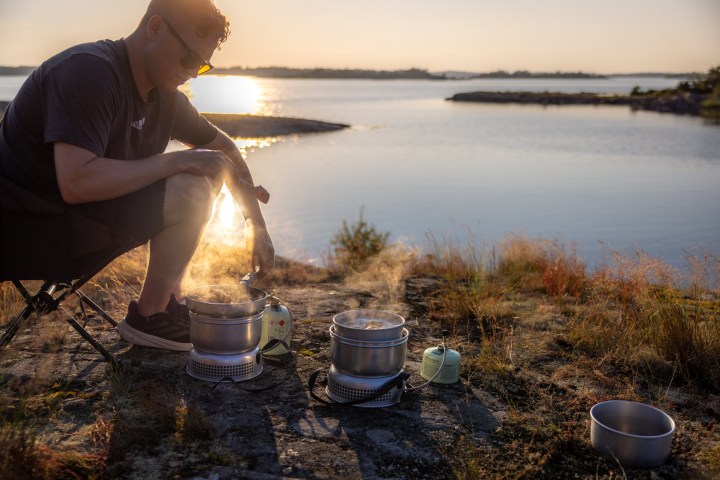Person cooking on portable stoves near a lake with sunset in the background.