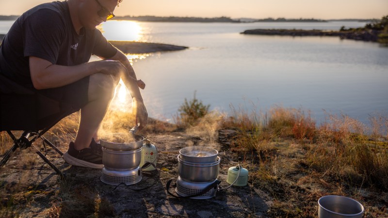 Person cooking on portable stoves near a lake with sunset in the background.