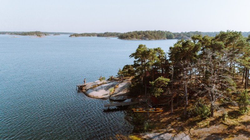 Person on rocky island with trees, surrounded by calm water and distant landmasses.