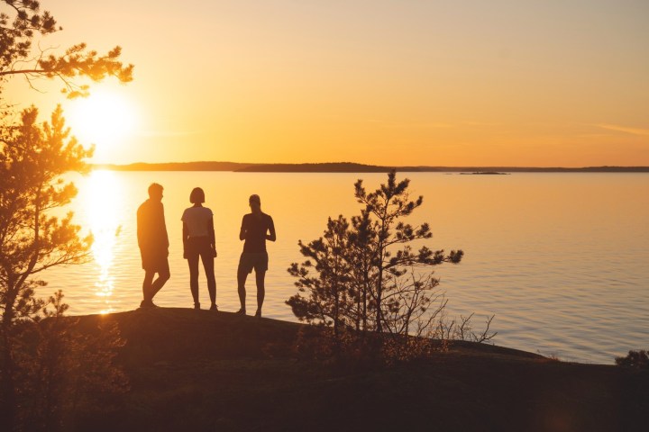 Silhouettes of three people standing near trees by a lake during sunset.