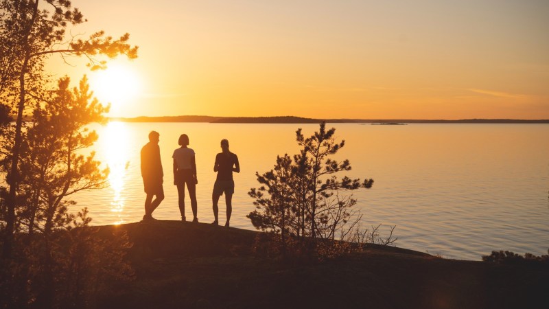 Silhouettes of three people standing near trees by a lake during sunset.