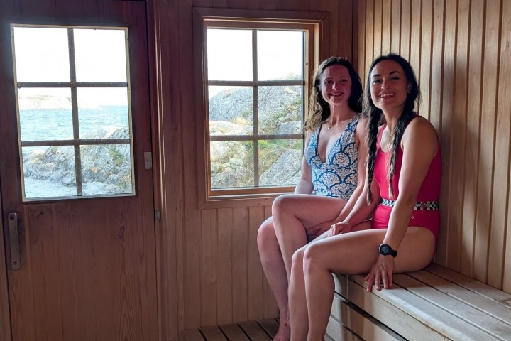 Two women in swimsuits sitting in a wooden sauna with a view of rocks and water through the windows.