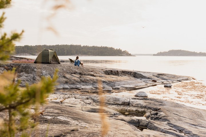 Person sitting near a tent on a rocky shore, overlooking a calm body of water at sunset.