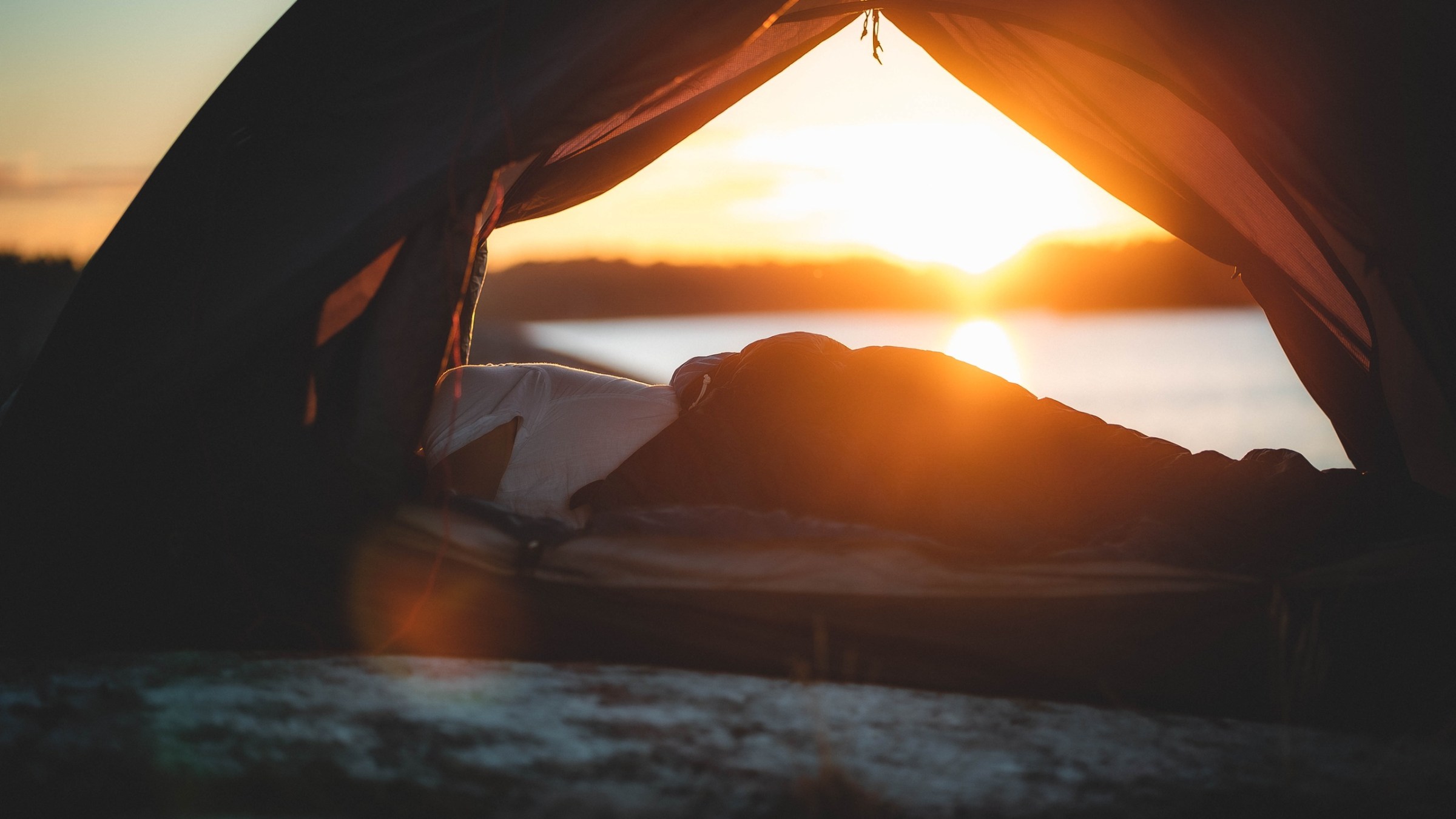 Person lying inside a tent, watching sunset by a lake.