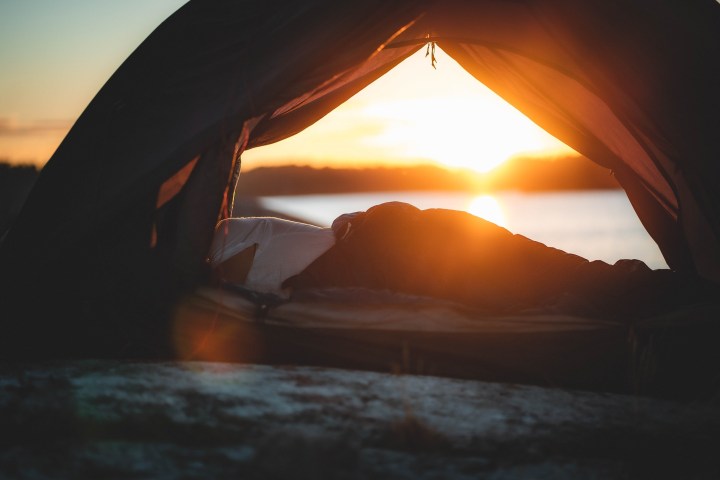 Person lying inside a tent, watching sunset by a lake.
