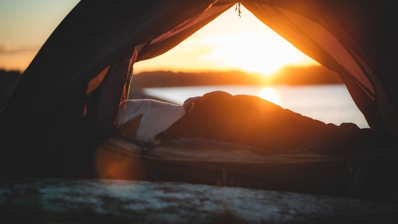 Person lying inside a tent, watching sunset by a lake.