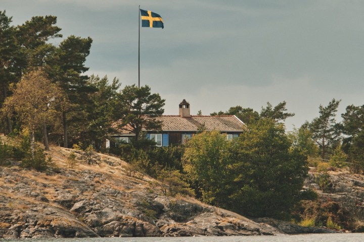 Swedish flag on a pole near a house surrounded by trees and rocky shoreline.