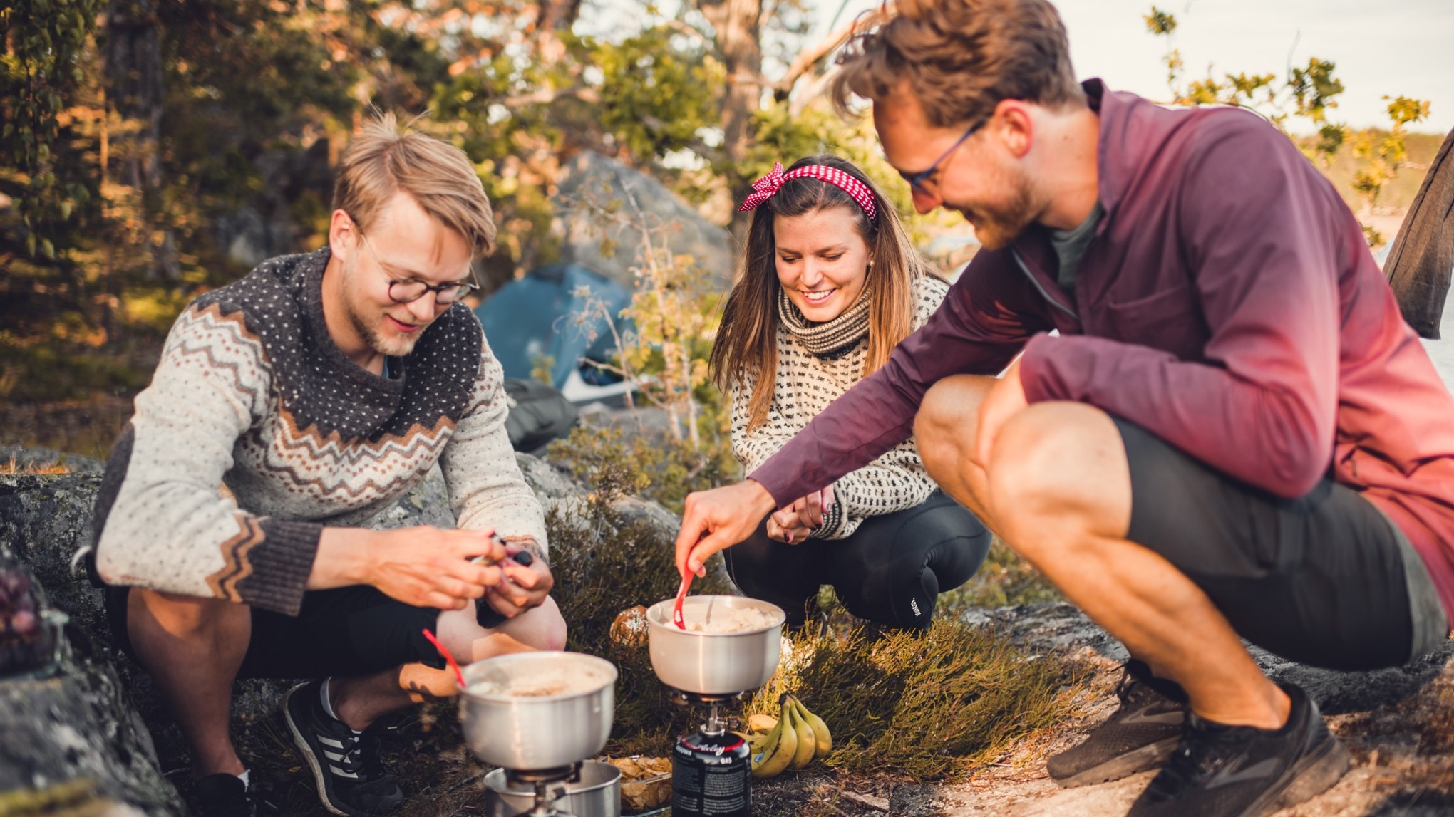 Three people cooking outdoors with camping gear, smiling and enjoying the activity.
