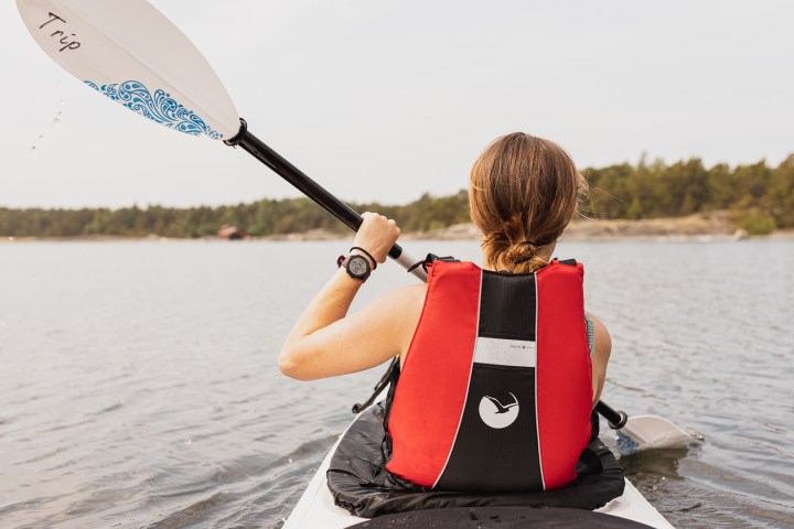 Person kayaking on calm water, wearing red life jacket, holding paddle, with forested shore in background.