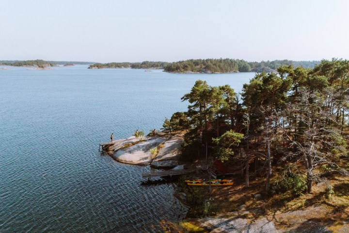 Aerial view of rocky shoreline with pine trees and kayaks; person standing on rock, calm water and islands in distance.