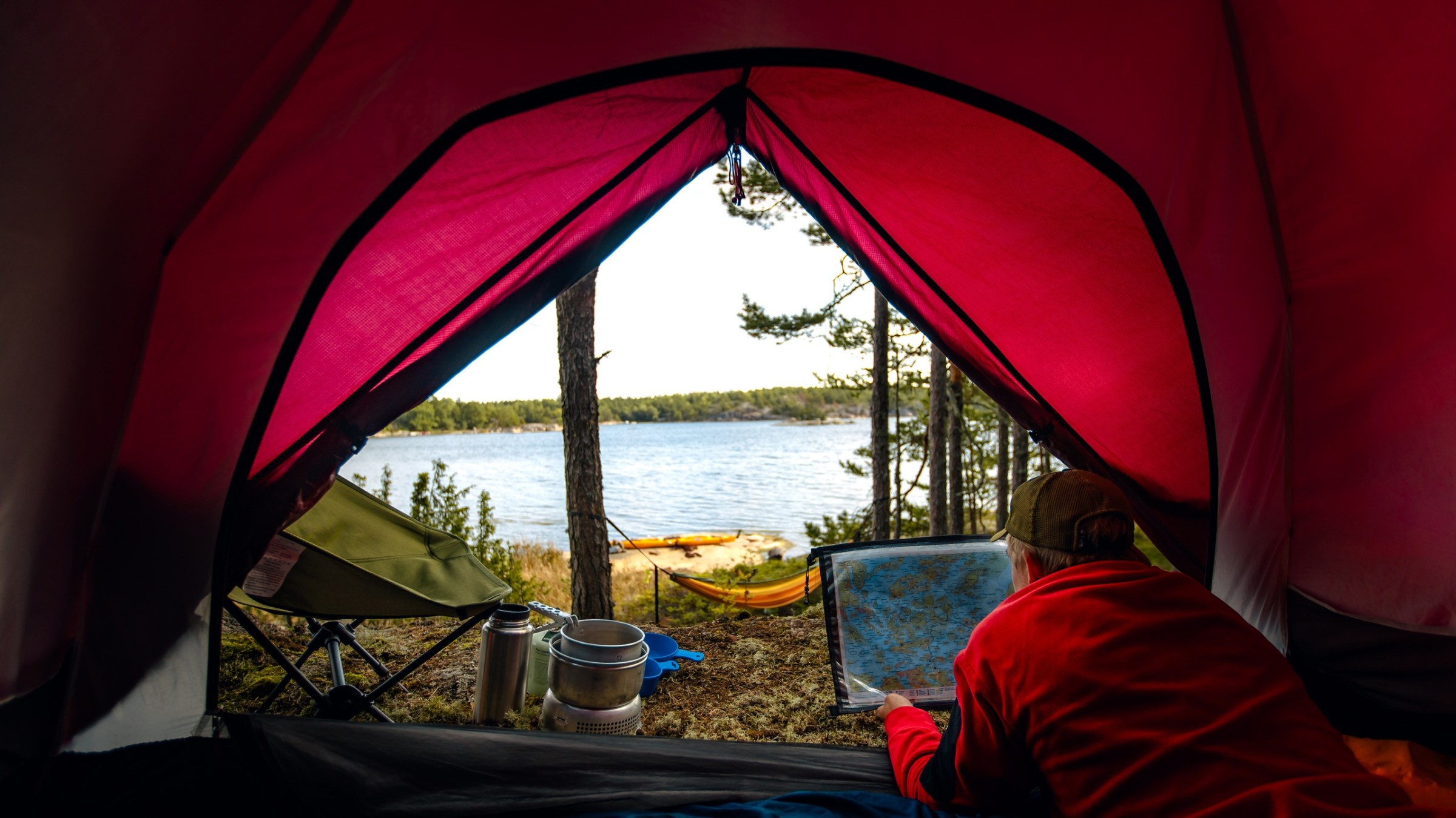 Person inside red tent looking at map, lake and forest view outside.