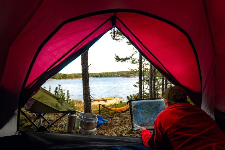 Person inside red tent looking at map, lake and forest view outside.