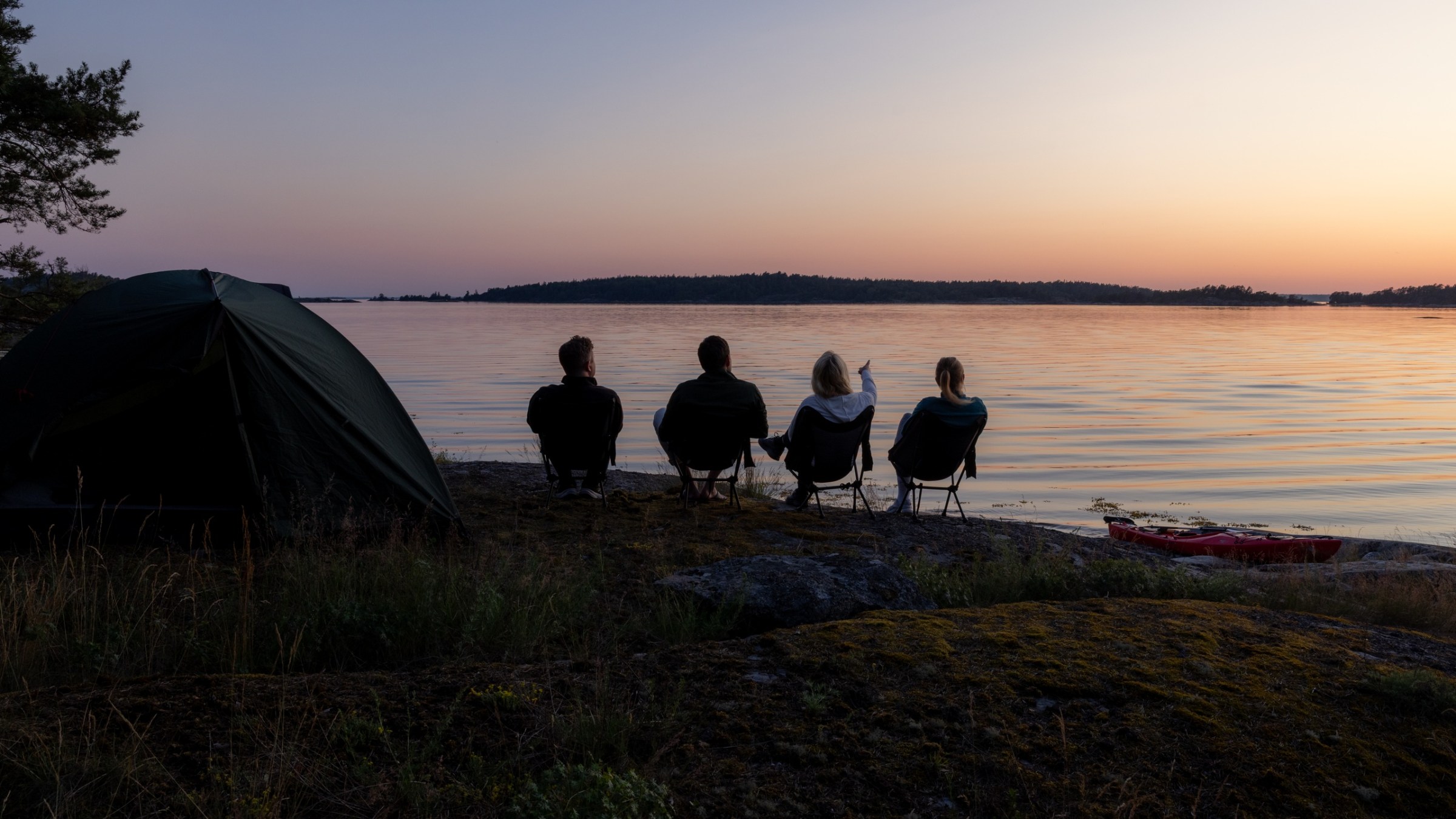 Four people seated by a tent on a lakeshore at sunset, looking out over calm waters.