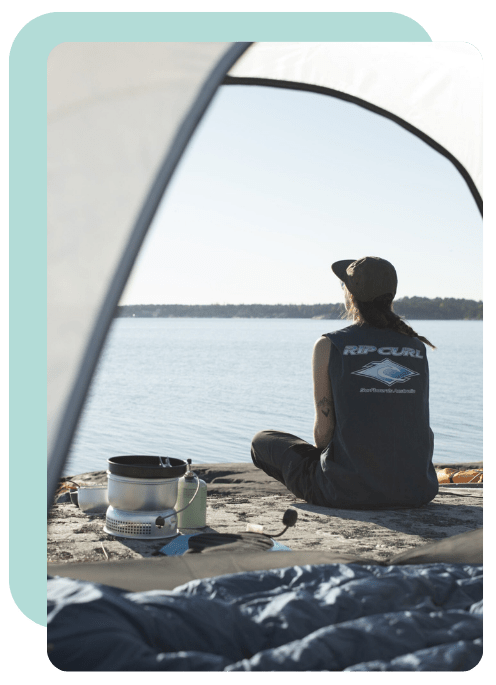 Person sitting by a lake, seen from inside a tent, with camping gear outside.