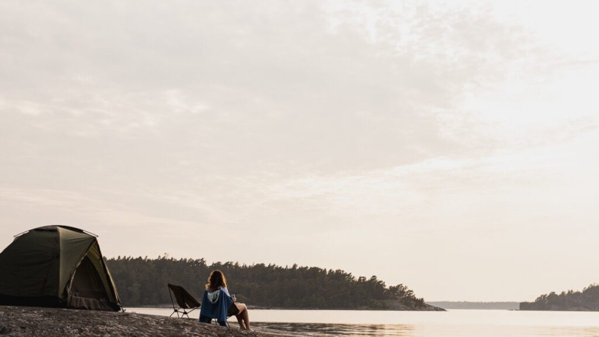 Person sitting near tent on rocky shore by calm water under cloudy sky.