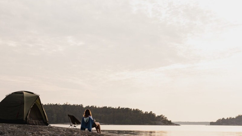 Person sitting near tent on rocky shore by calm water under cloudy sky.