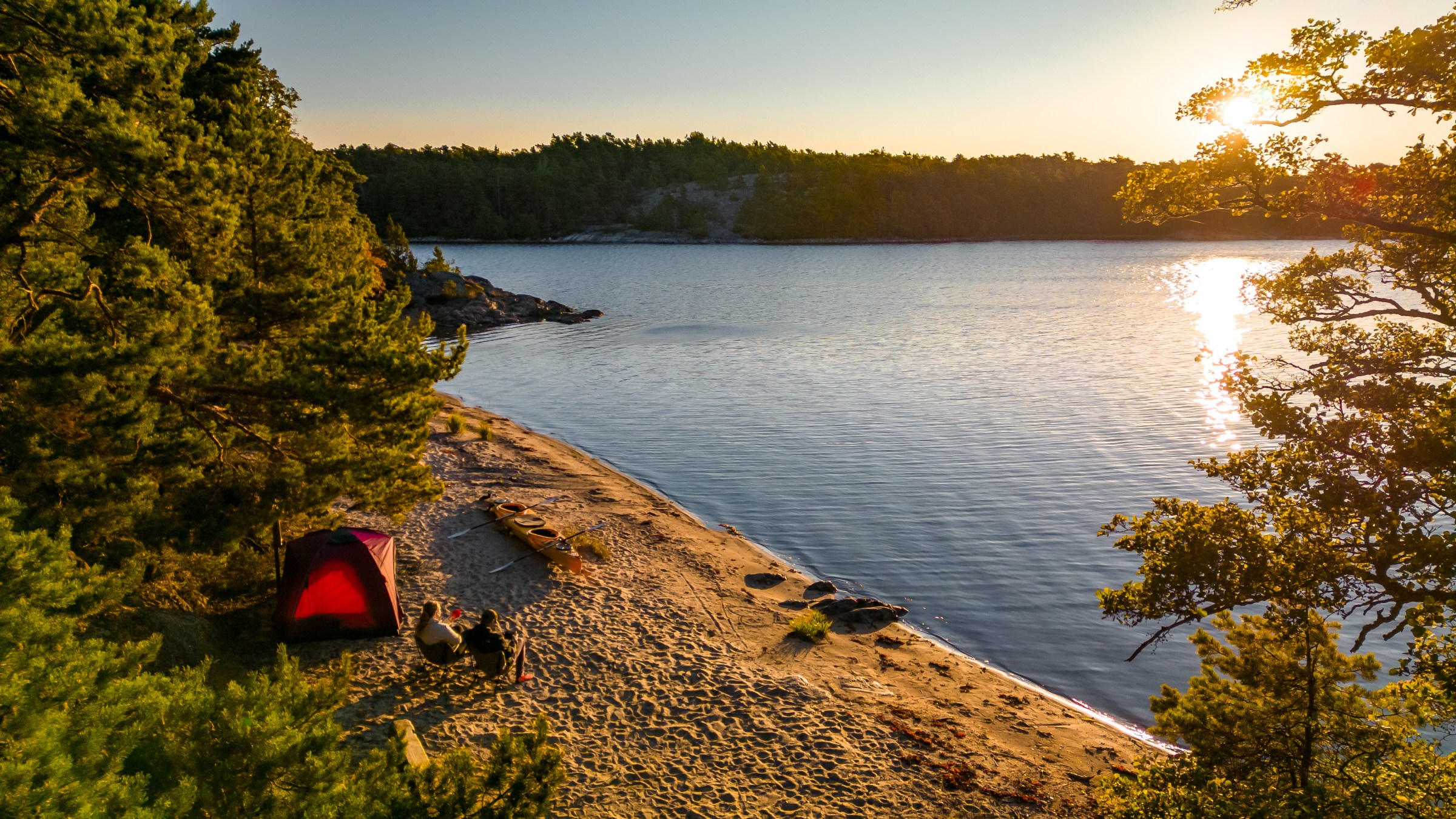 Tent and campsite on a lake shore surrounded by trees at sunset.