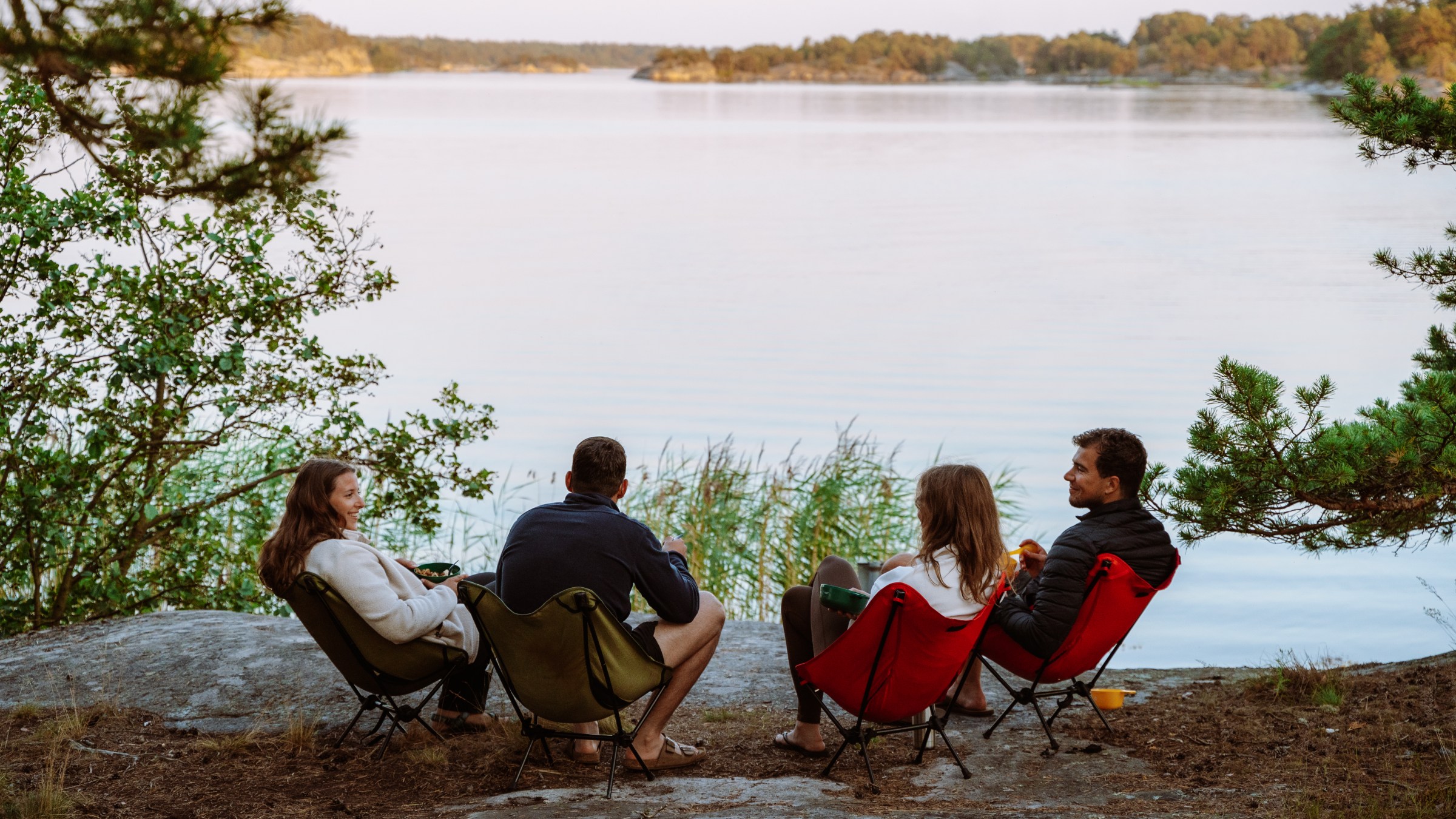 Four people seated on chairs by a lake, surrounded by trees, at sunset.