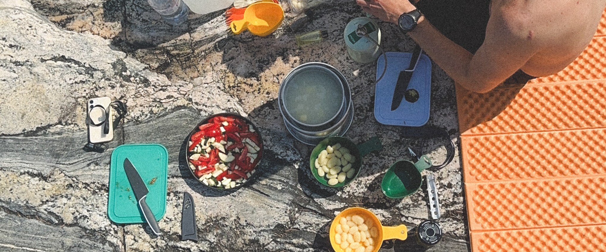Two people eating on rocky terrain with camping gear and food items.